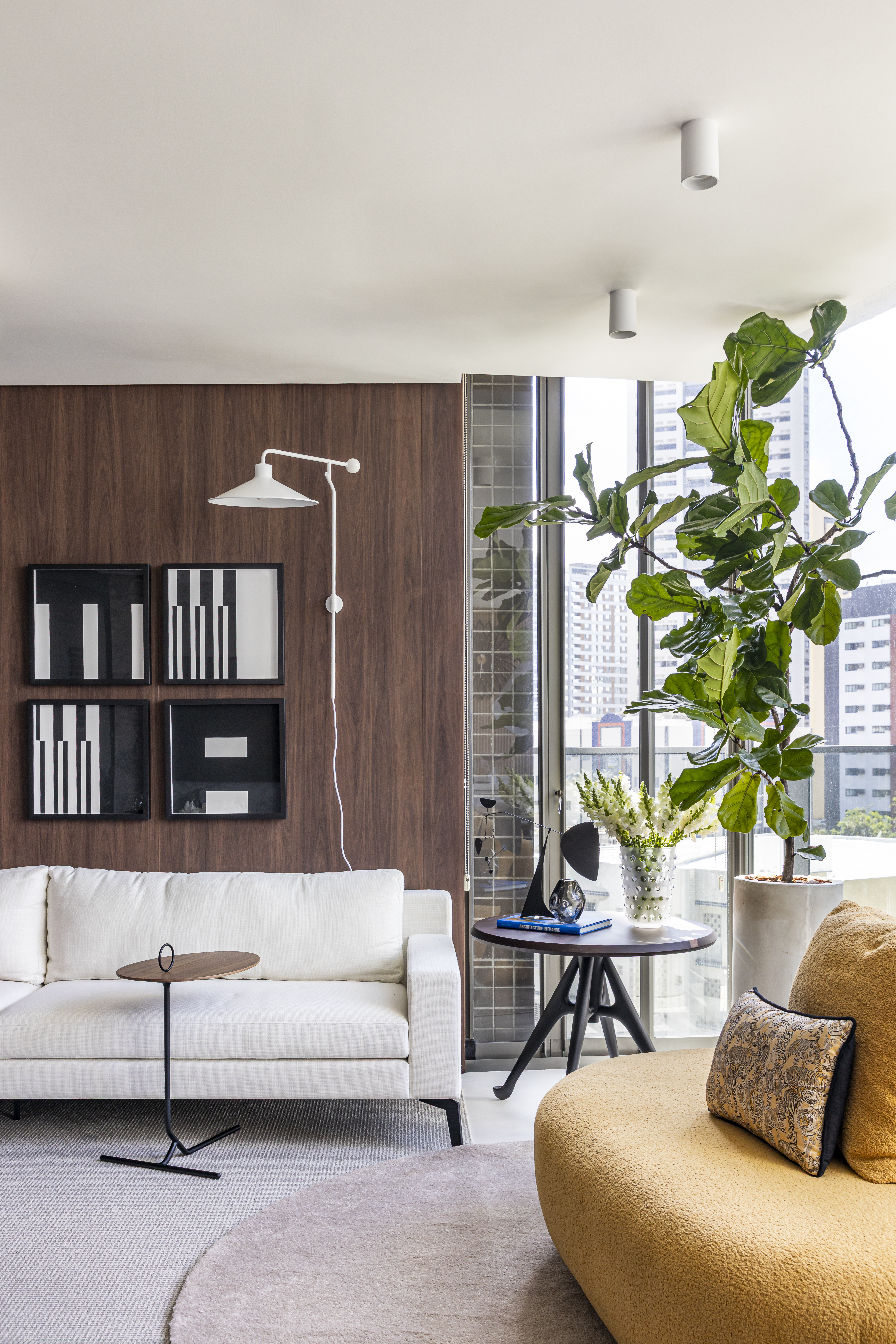 Apartamento em Recife &eacute; transformado em ref&uacute;gio urbano sofisticado e atemporal. Projeto de Rom&aacute;rio Rodrigues Arquitetura. Na foto, Sala de estar moderna com sof&aacute; branco, mesa lateral redonda de madeira e metal, e um tapete cinza claro. Na parede de madeira escura, quatro quadros abstratos em preto e branco e um arandela branca. &Agrave; direita, uma poltrona amarela texturizada, uma mesa de centro redonda com vaso de flores brancas, livros e objetos decorativos. Uma grande planta verde em vaso de cimento est&aacute; perto da janela, revelando pr&eacute;dios ao fundo