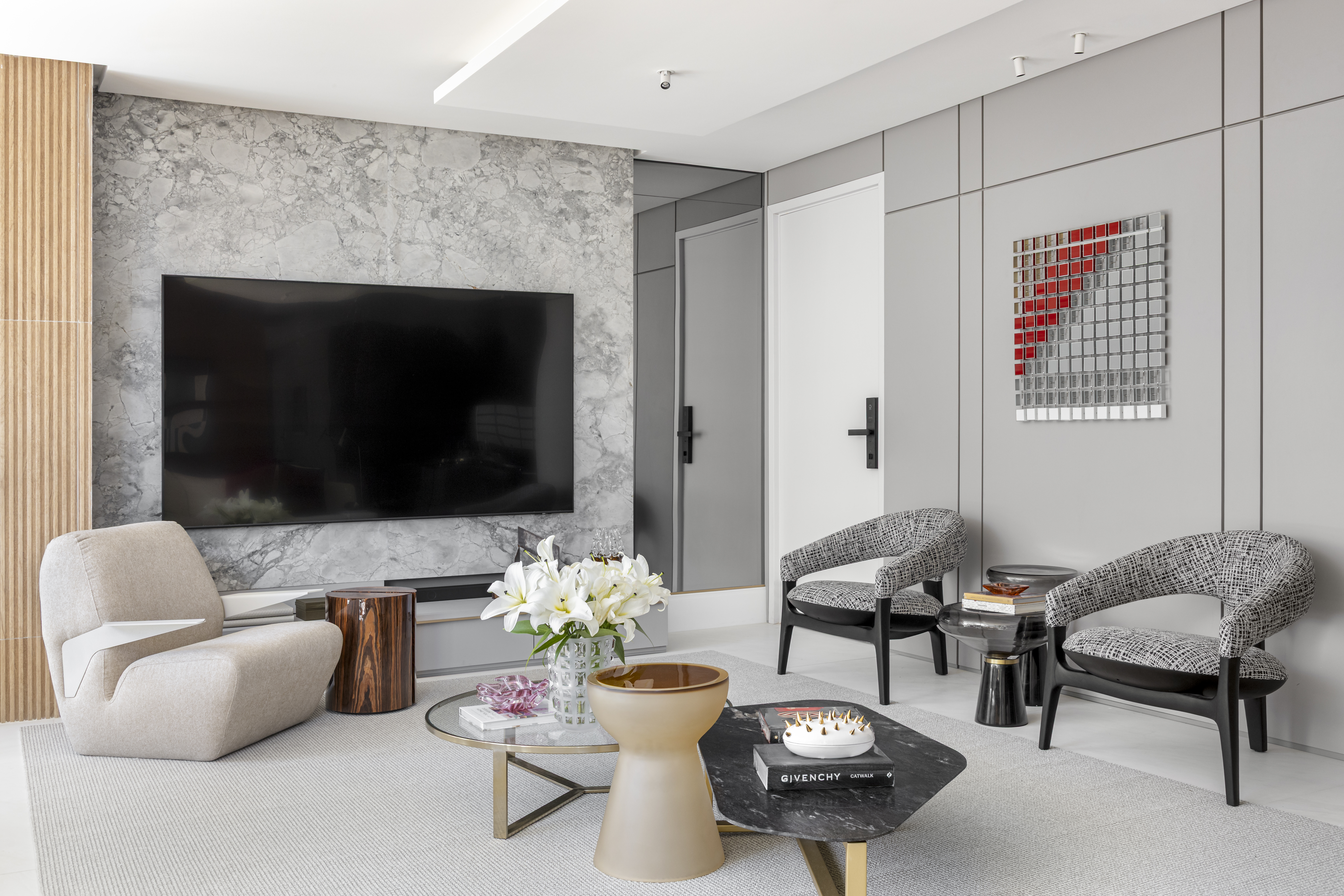 Apartamento em Recife &eacute; transformado em ref&uacute;gio urbano sofisticado e atemporal. Projeto de Rom&aacute;rio Rodrigues Arquitetura. Na foto, Sala de estar moderna com paredes cinzas e uma TV grande sobre um painel de m&aacute;rmore. H&aacute; um sof&aacute; bege, duas poltronas estampadas, mesas de centro com livros e um vaso de l&iacute;rios brancos. Uma obra de arte com quadrados vermelhos e espelhados decora a parede.