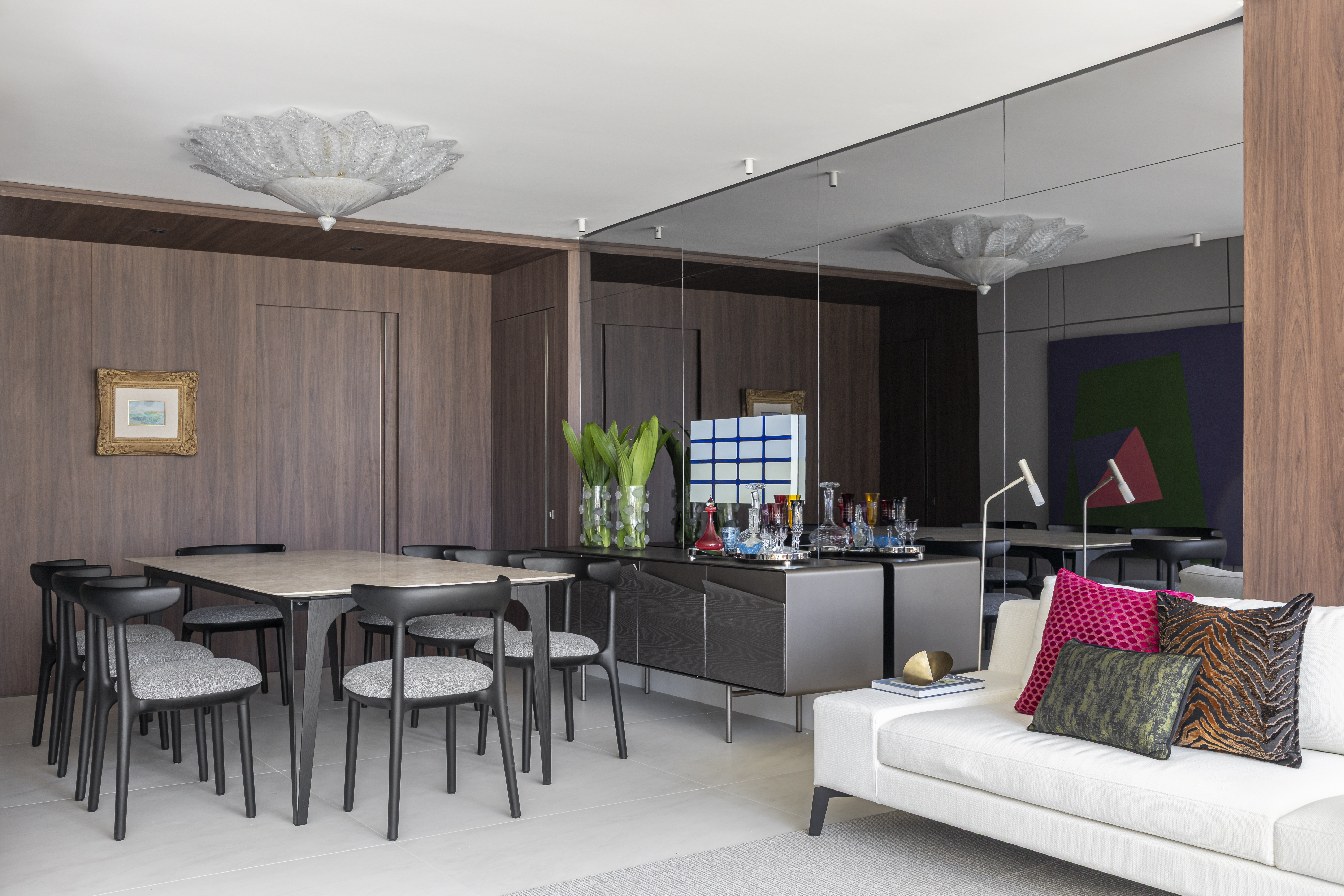 Apartamento em Recife &eacute; transformado em ref&uacute;gio urbano sofisticado e atemporal. Projeto de Rom&aacute;rio Rodrigues Arquitetura. Na foto, Sala de jantar e estar integradas com mesa retangular de tampo claro e oito cadeiras pretas com assento cinza. Ao fundo, painel de madeira escura com um quadro pequeno e um aparador cinza com vasos de plantas e copos coloridos. &Agrave; direita, um sof&aacute; branco com almofadas coloridas e um espelho grande refletindo uma obra de arte abstrata