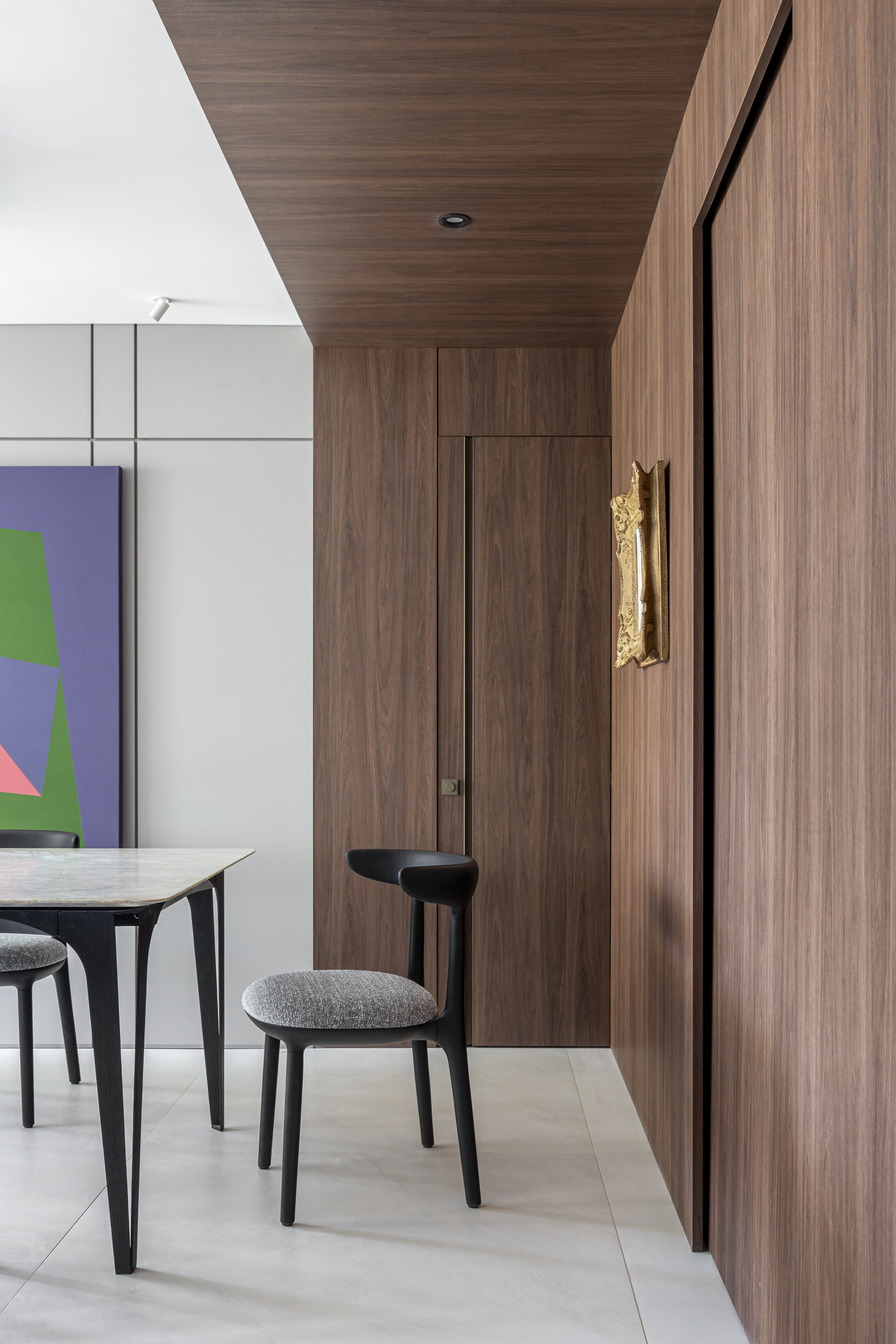 Apartamento em Recife &eacute; transformado em ref&uacute;gio urbano sofisticado e atemporal. Projeto de Rom&aacute;rio Rodrigues Arquitetura. Na foto, Ambiente interno com mesa de jantar de tampo claro e cadeiras pretas com assento cinza, ao lado de uma parede e porta de madeira escura. No fundo, uma obra de arte abstrata com formas geom&eacute;tricas em verde, roxo e rosa. Uma lumin&aacute;ria dourada e irregular decora a parede de madeira
