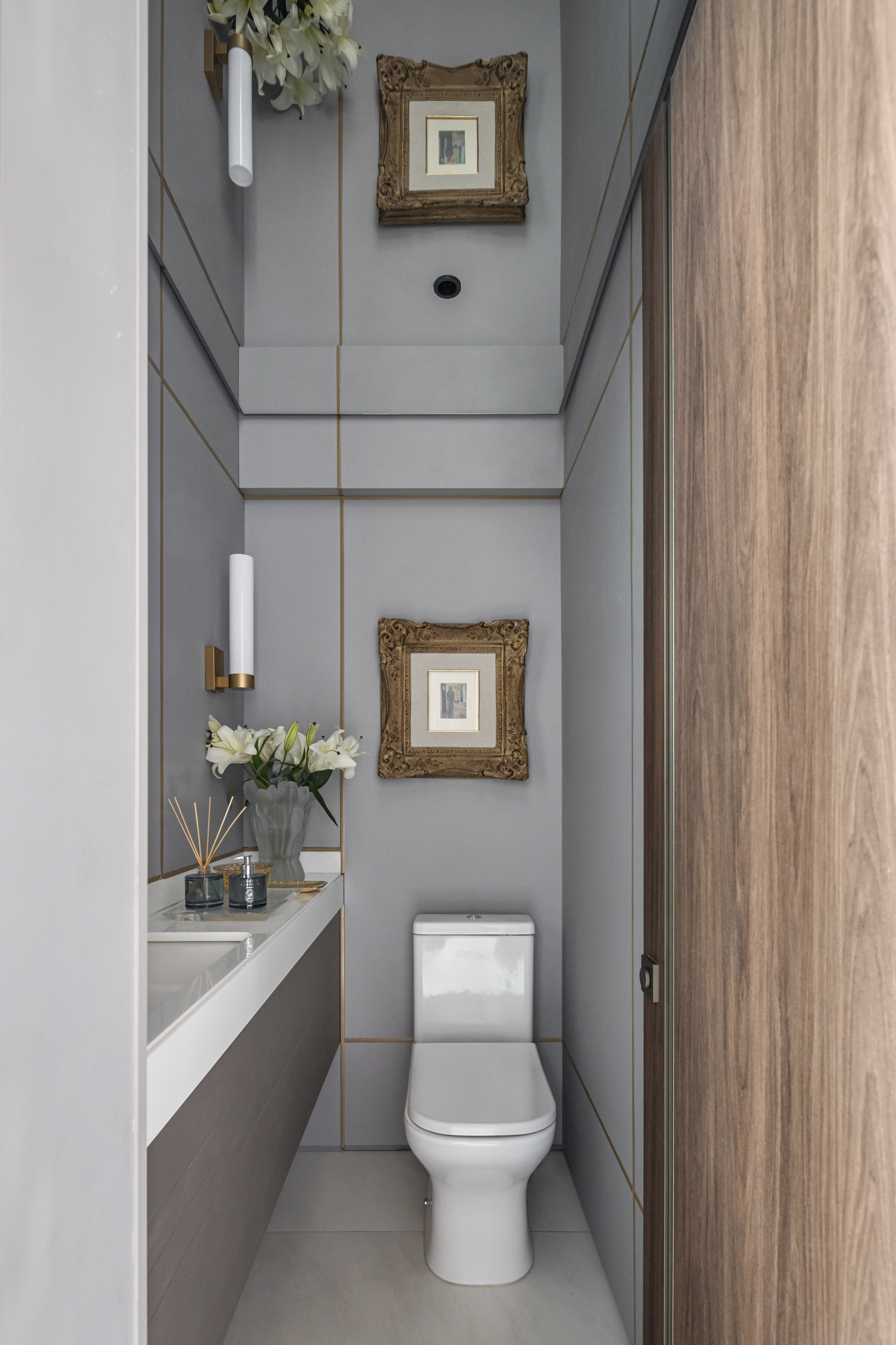 Apartamento em Recife &eacute; transformado em ref&uacute;gio urbano sofisticado e atemporal. Projeto de Rom&aacute;rio Rodrigues Arquitetura. Na foto, Lavabo moderno com paredes cinza e detalhes dourados, pia com bancada branca, vaso sanit&aacute;rio branco e porta de madeira. H&aacute; dois quadros dourados e arandelas brancas nas paredes, al&eacute;m de um vaso com l&iacute;rios brancos e difusores de ambiente sobre a pia