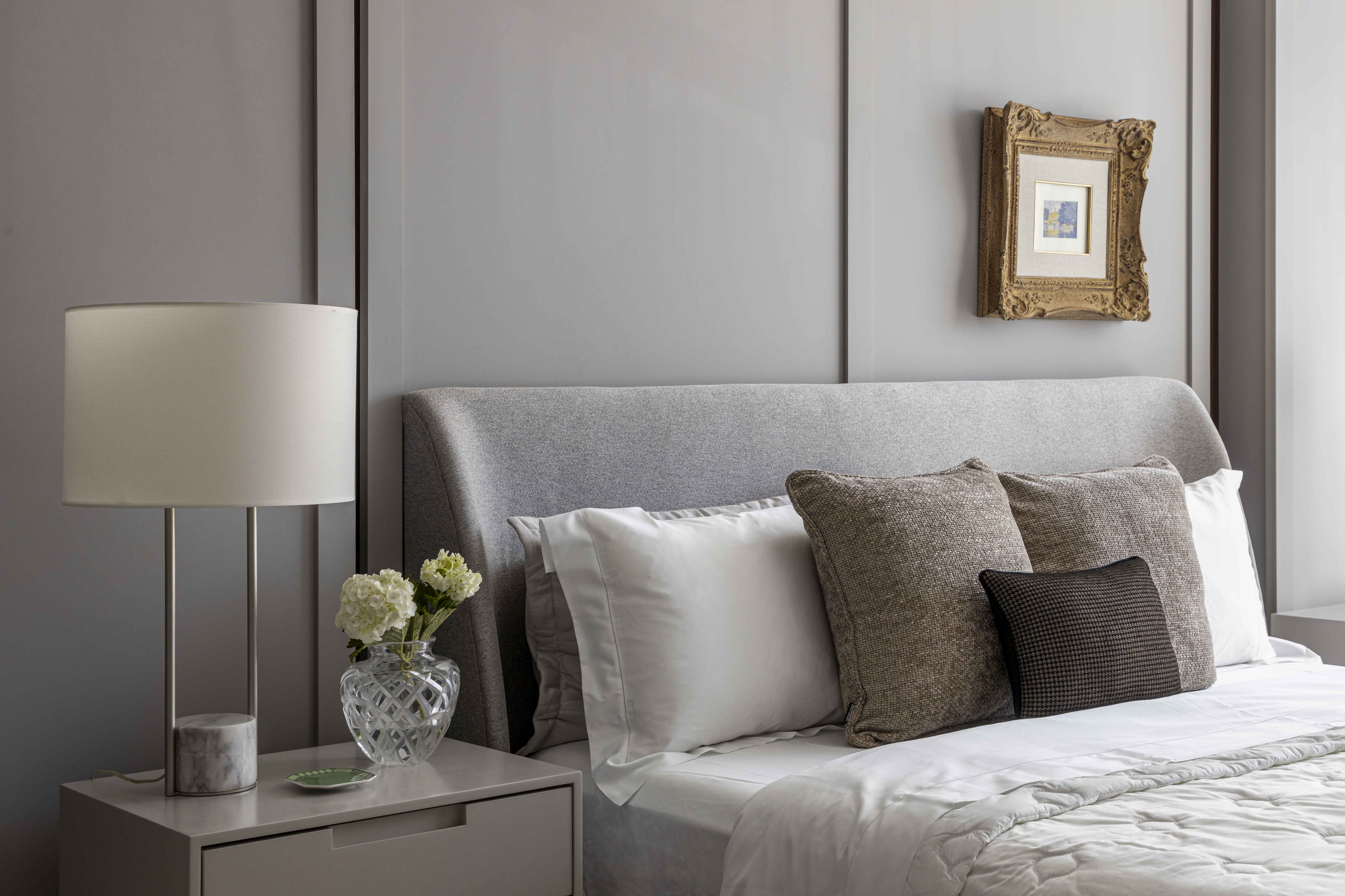 Apartamento em Recife &eacute; transformado em ref&uacute;gio urbano sofisticado e atemporal. Projeto de Rom&aacute;rio Rodrigues Arquitetura. Na foto, Quarto com cama cinza, cabeceira estofada, len&ccedil;&oacute;is brancos e almofadas marrons. Na mesa de cabeceira, um abajur com base de m&aacute;rmore, vaso de cristal com flores brancas e um pequeno prato verde. Na parede cinza, um quadro com moldura dourada ornamentada