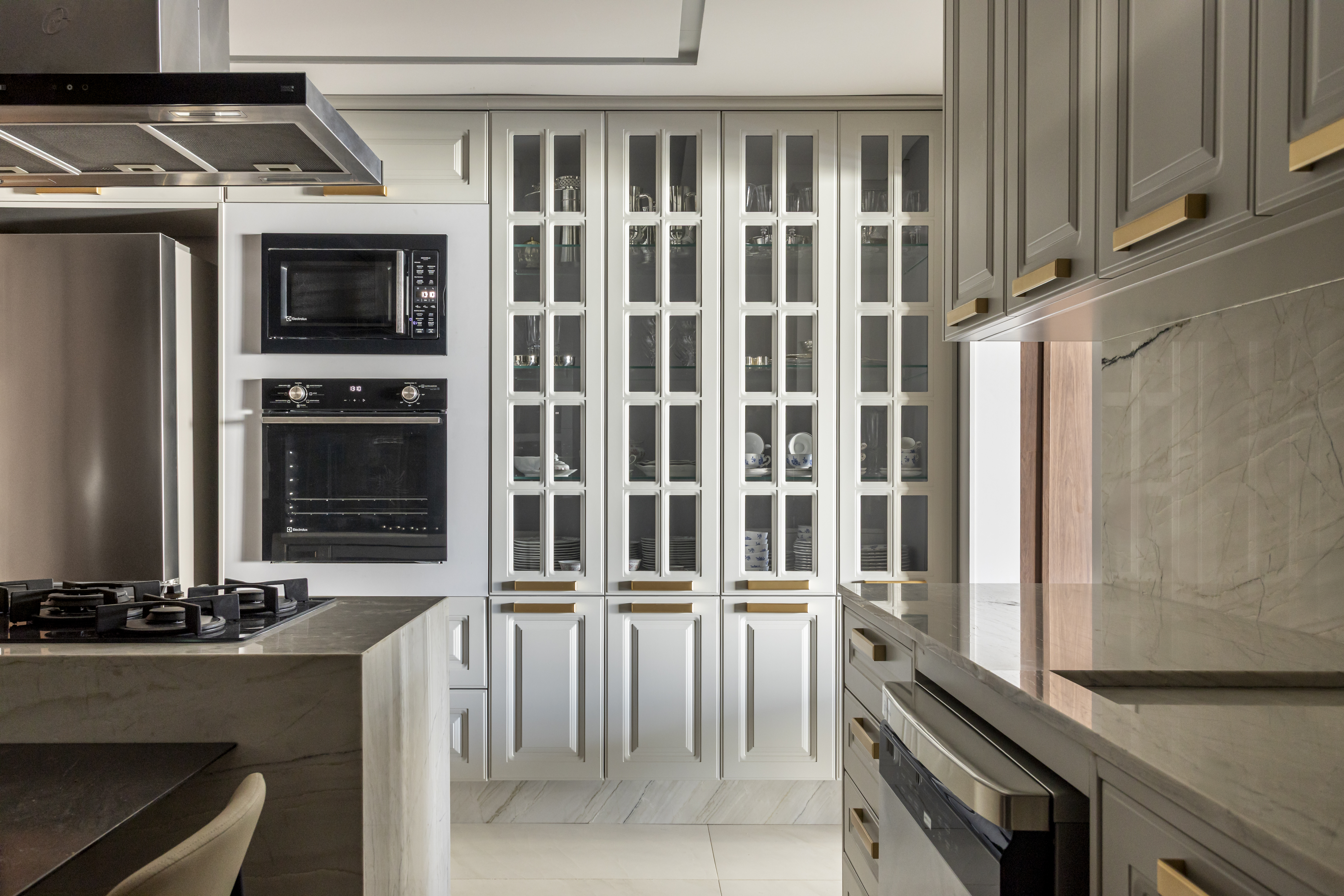 Apartamento em Recife &eacute; transformado em ref&uacute;gio urbano sofisticado e atemporal. Projeto de Rom&aacute;rio Rodrigues Arquitetura. Na foto, Cozinha moderna com arm&aacute;rios brancos e cinzas, bancadas de m&aacute;rmore claro e eletrodom&eacute;sticos embutidos. &Agrave; esquerda, geladeira, micro-ondas e forno pretos. Ao centro, arm&aacute;rios superiores com portas de vidro e pratos empilhados. &Agrave; direita, arm&aacute;rios cinzas com puxadores dourados e bancada de m&aacute;rmore. O ch&atilde;o &eacute; de m&aacute;rmore branco.