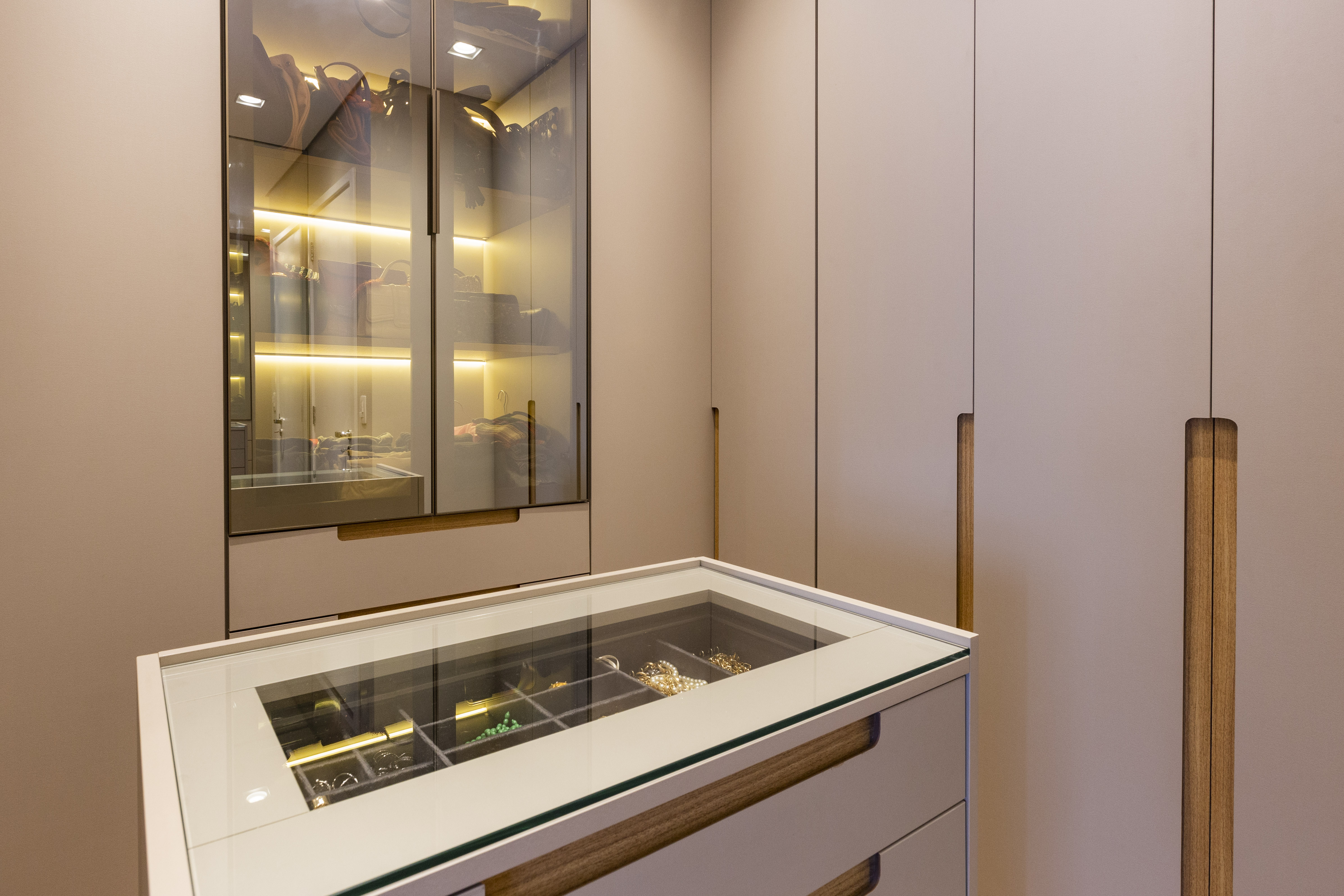 Projetos com vidro reflecta, o material que &eacute; espelhado e transparente ao mesmo tempo. Projeto de Daniela Funari. Na foto, Um closet moderno com arm&aacute;rios bege e puxadores de madeira. &Agrave; esquerda, um arm&aacute;rio com portas de vidro transparente e ilumina&ccedil;&atilde;o interna amarela exibe bolsas e sapatos. No centro, uma c&ocirc;moda baixa com tampo de vidro revela joias e acess&oacute;rios organizados em divis&oacute;rias.