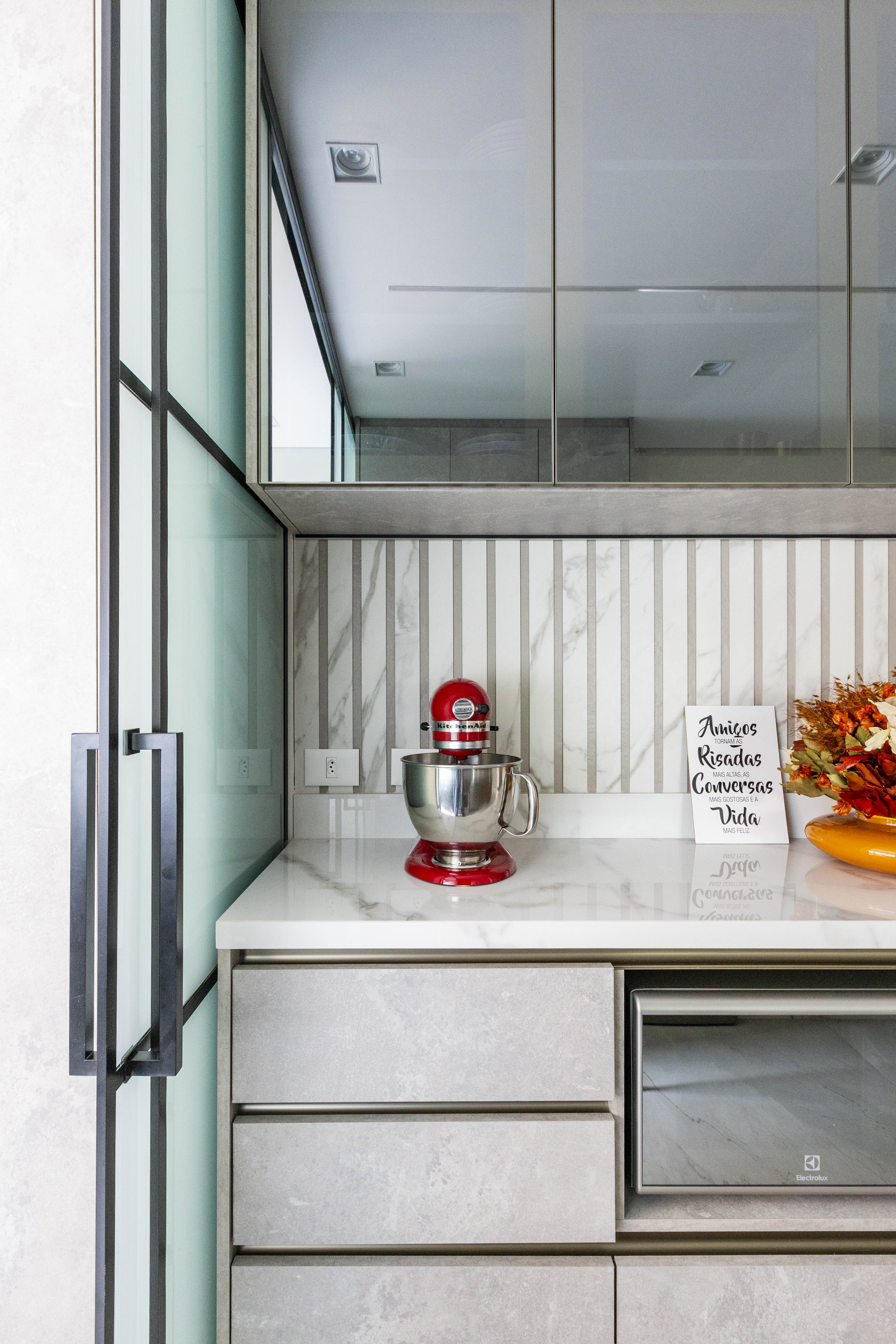 Projetos com vidro reflecta, o material que &eacute; espelhado e transparente ao mesmo tempo. Projeto de Daniela Funari. Na foto, Cozinha moderna com bancada de m&aacute;rmore branco, batedeira vermelha, painel decorativo com listras de m&aacute;rmore e frase Amigos, Risadas, Conversas, Vida. Arm&aacute;rios cinza e forno de micro-ondas embutido, com arm&aacute;rios superiores espelhados e porta de vidro canelado &agrave; esquerda