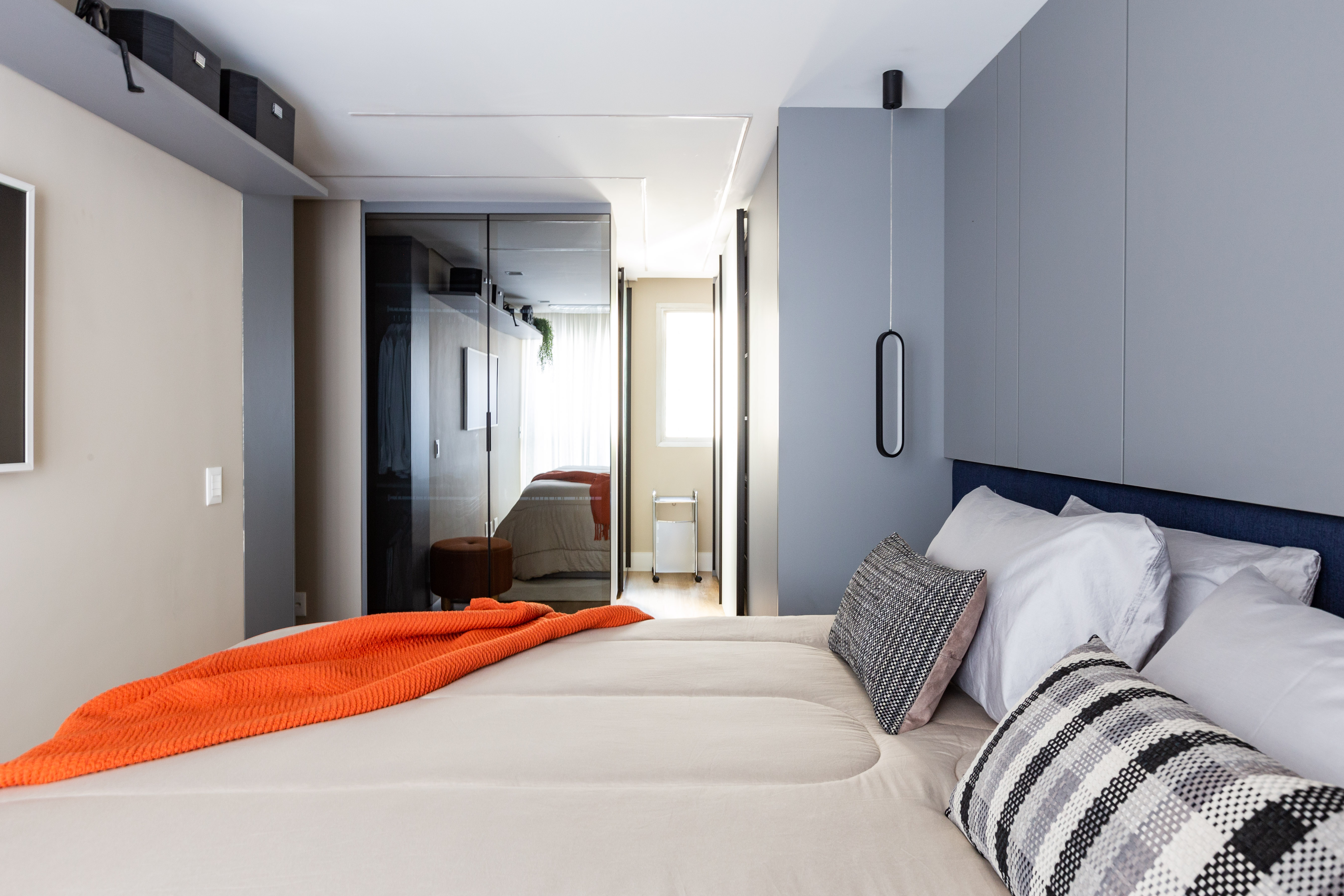 Projetos com vidro reflecta, o material que &eacute; espelhado e transparente ao mesmo tempo. Projeto de Daniela Funari. Na foto, Quarto moderno com cama de casal bege, manta laranja dobrada, almofadas brancas e estampadas. Parede cinza com lumin&aacute;ria pendente preta, arm&aacute;rio espelhado e prateleira superior com caixas escuras. Ao fundo, outro quarto com cama e janela iluminada
