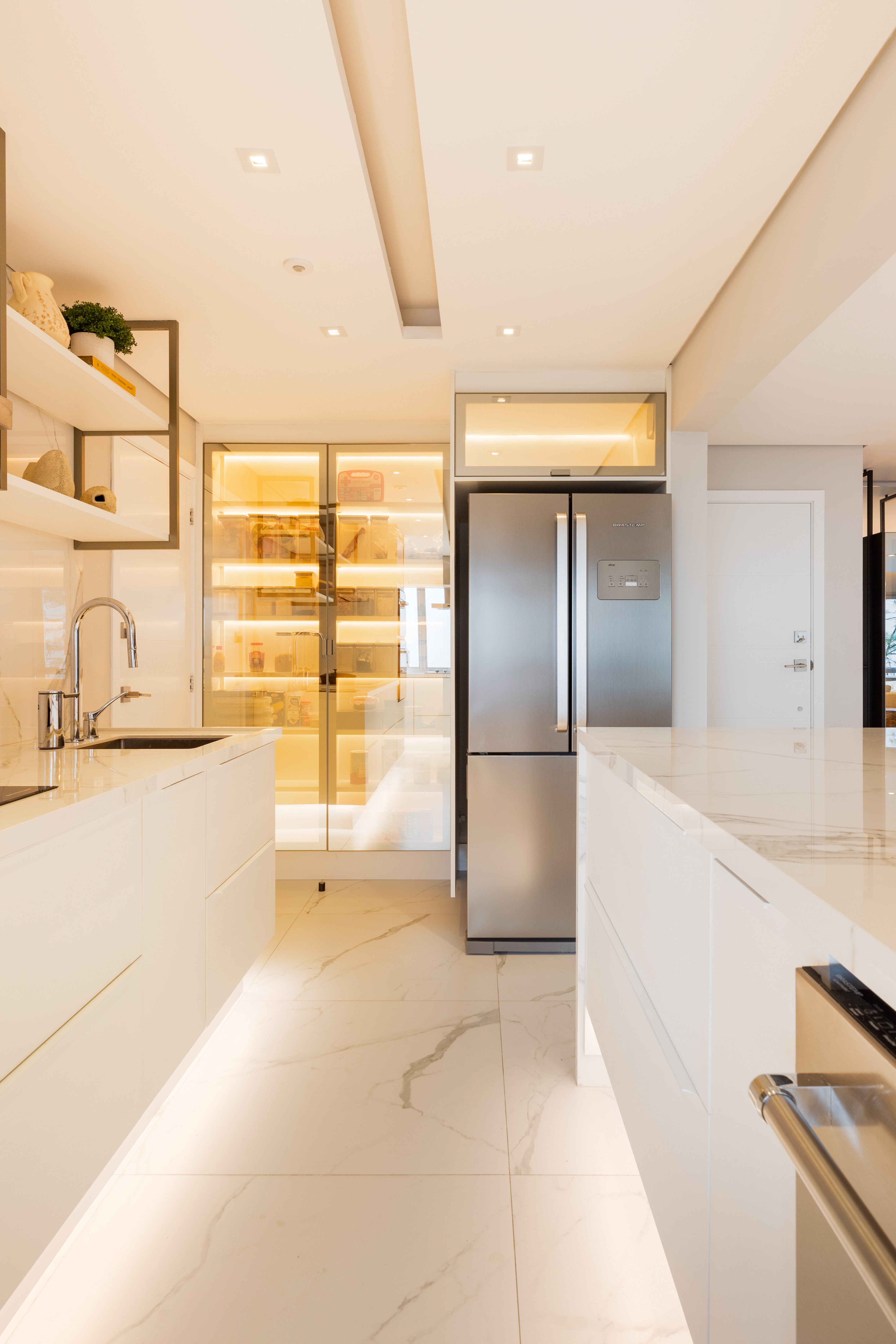 Projetos com vidro reflecta, o material que &eacute; espelhado e transparente ao mesmo tempo. Projeto de Daniela Funari. Na foto, Cozinha moderna com arm&aacute;rios brancos, bancadas de m&aacute;rmore e piso de porcelanato branco com veios cinzas. &Agrave; esquerda, pia com torneira cromada e prateleiras decoradas. Ao fundo, despensa com portas de vidro e ilumina&ccedil;&atilde;o interna amarela. &Agrave; direita, geladeira de a&ccedil;o inoxid&aacute;vel e uma ilha com detalhes met&aacute;licos. O ambiente &eacute; bem iluminado com luzes embutidas no teto e sob os arm&aacute;rios